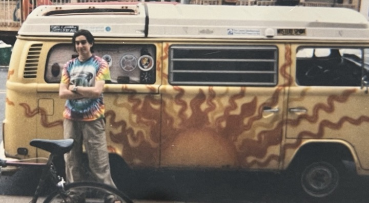 Young Jake in a tie-dye shirt, arms crossed, in front of a flame-painted VW bus